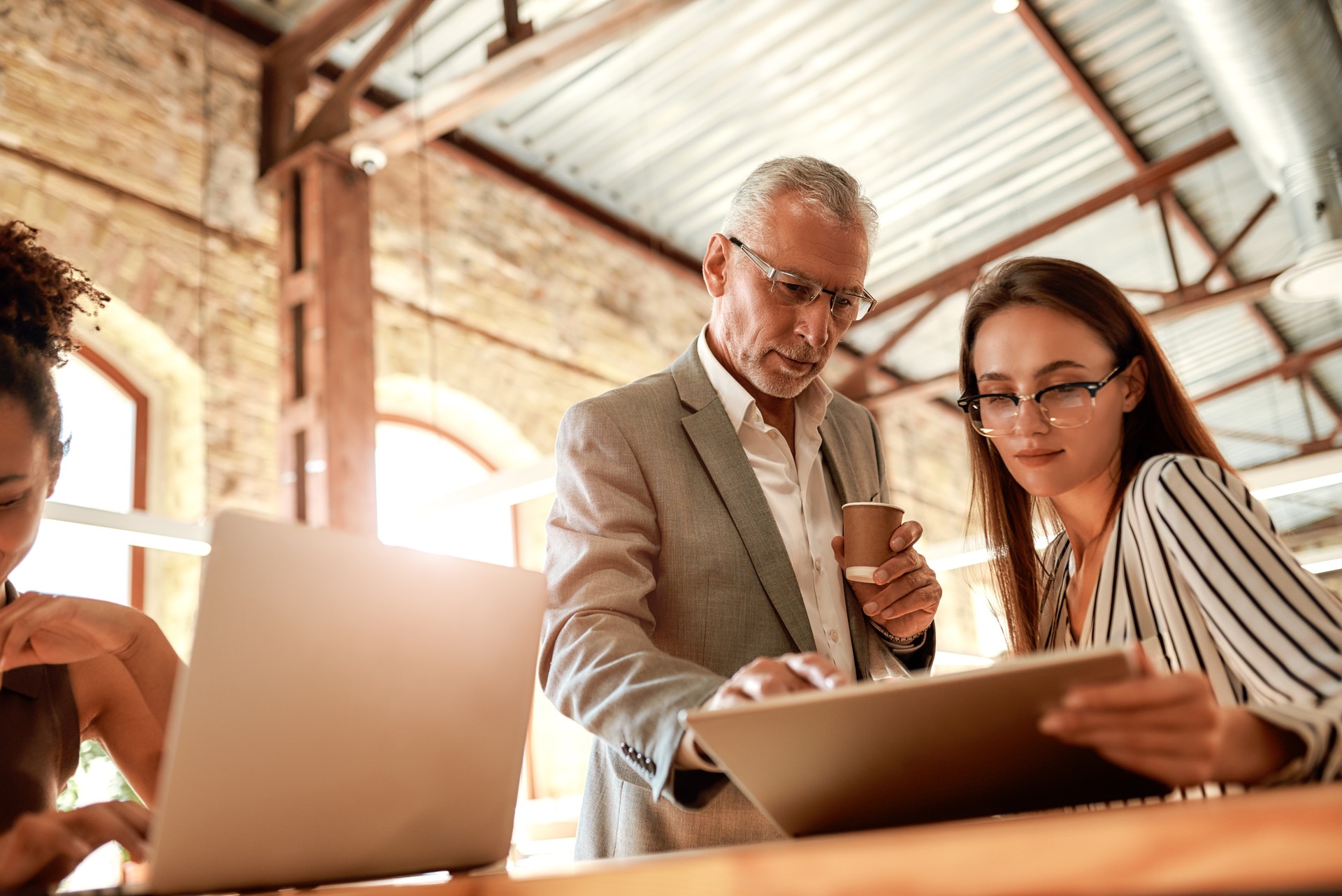 Sharing experience. Senior man in formal wear holding coffee cup and explaining something to his young female colleague while working together in the modern office