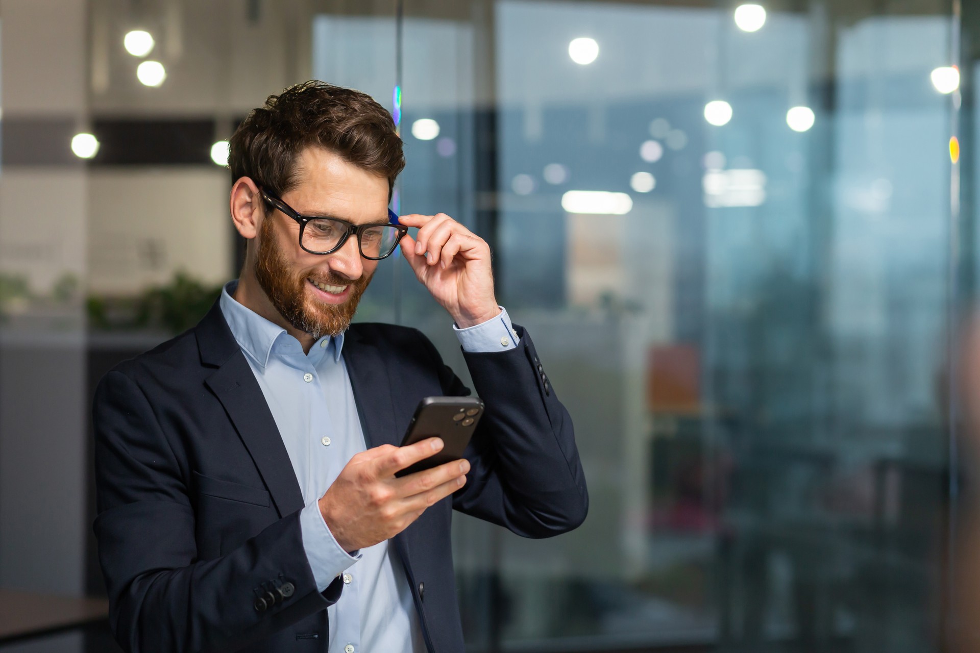 Successful financier investor works inside office at work, businessman in business suit uses telephone near window, man smiles and reads good news online from smartphone